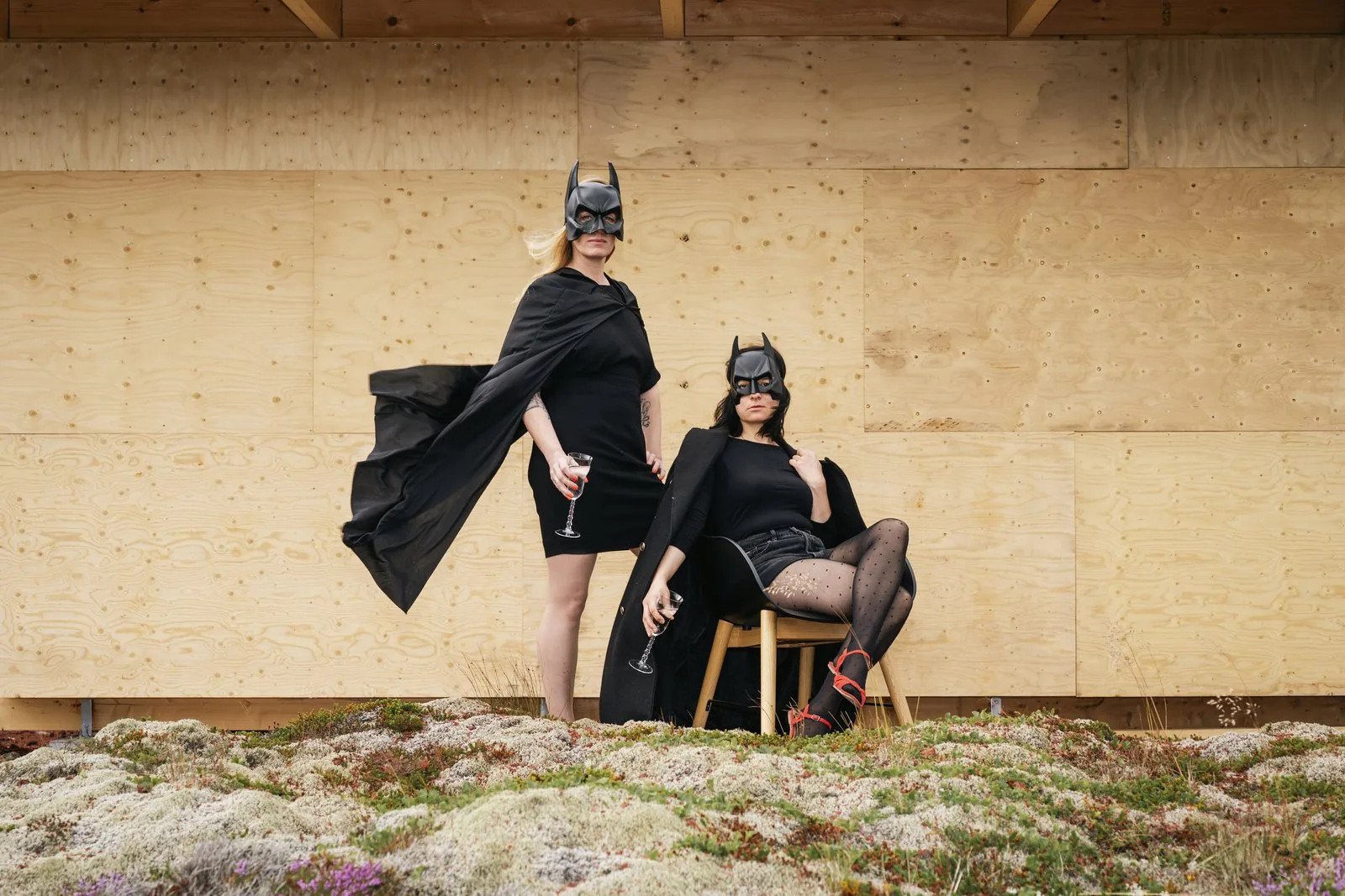 Anne und Sibille in Batman-Masken und Umhängen vor einer Holzwand, jede mit einem Glas in der Hand
