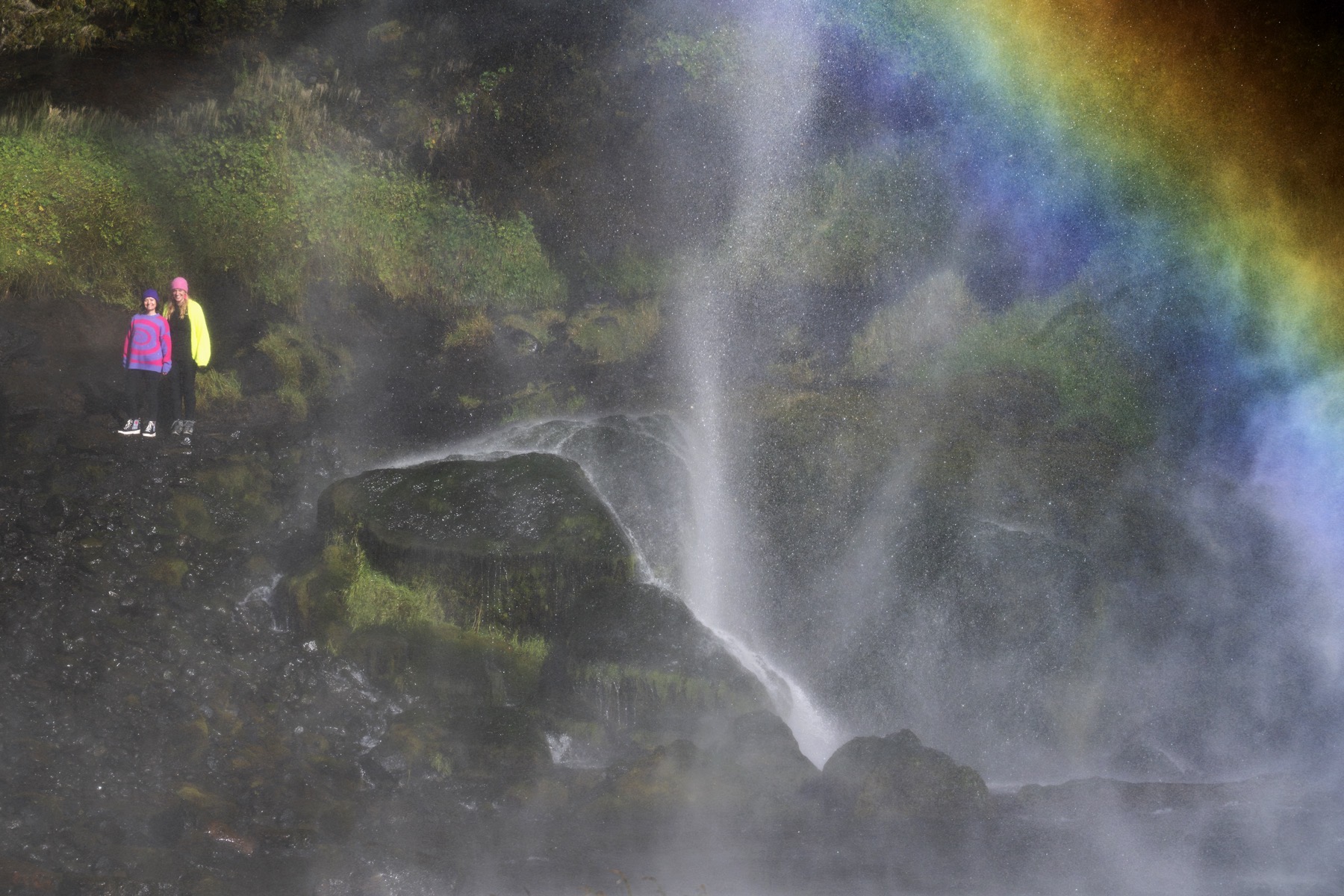 Anne und Sibille stehen klein neben einem isländischen Wasserfall, über ihnen ein Regenbogen
