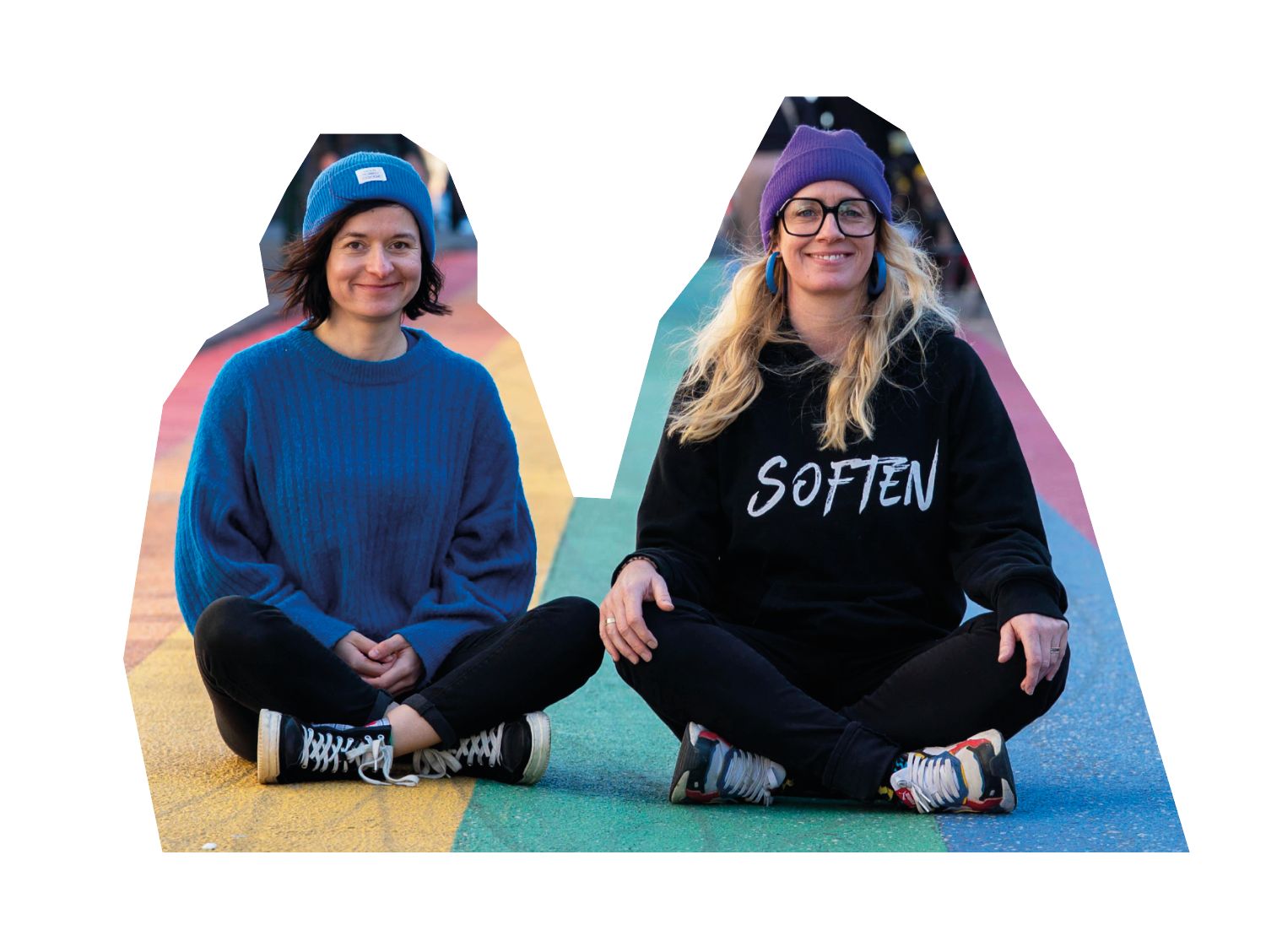 Anne und Sibille sitzen im Schneidersitz auf einer Regenbogenstraße, Sibille im Pullover mit der Aufschrift Soften