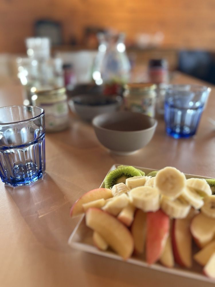 Eine Platte mit aufgeschnittener Kiwi, Banane und Apfel im Vordergrund, dahinter der gedeckte Frühstückstisch