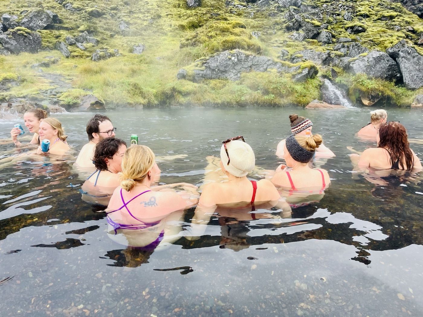 Eine Gruppe Frauen sitzt nebeneinander in einer heißen Quelle zwischen moosbewachsenen Felsen