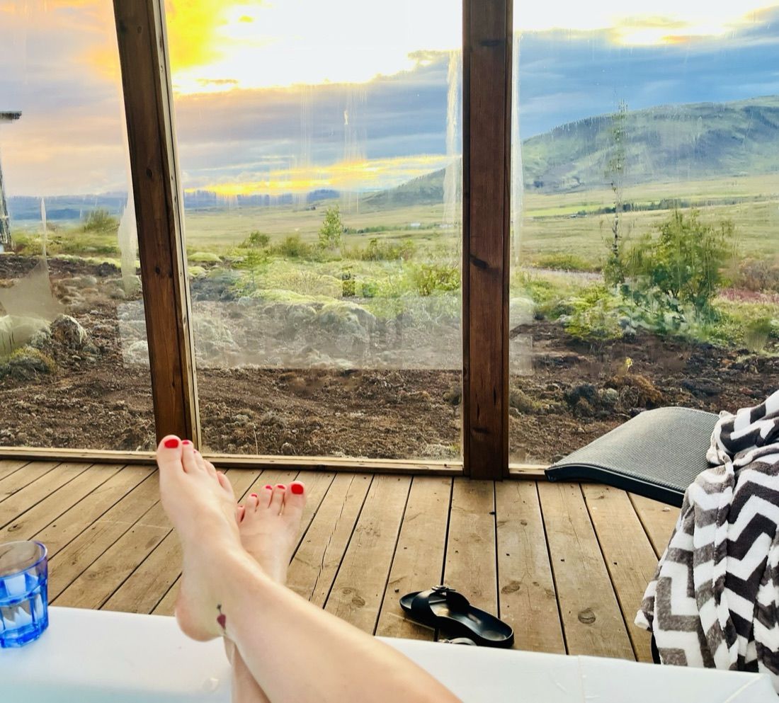 Blick aus dem Hot Tub über die Holzterrasse durch die Verglasung auf die Ebene, dahinter geht die Sonne unter