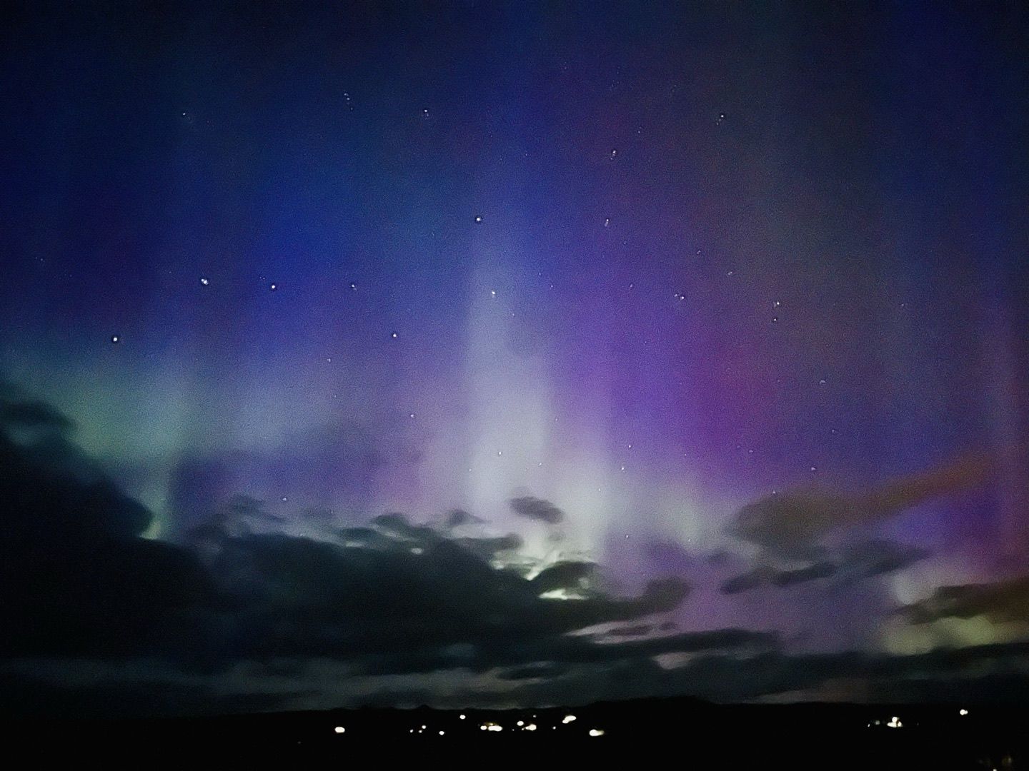 Polarlicht über der isländischen Ebene: grüne und violette Schleier über einer Wolkenbank, darunter die Lichter eines Ortes