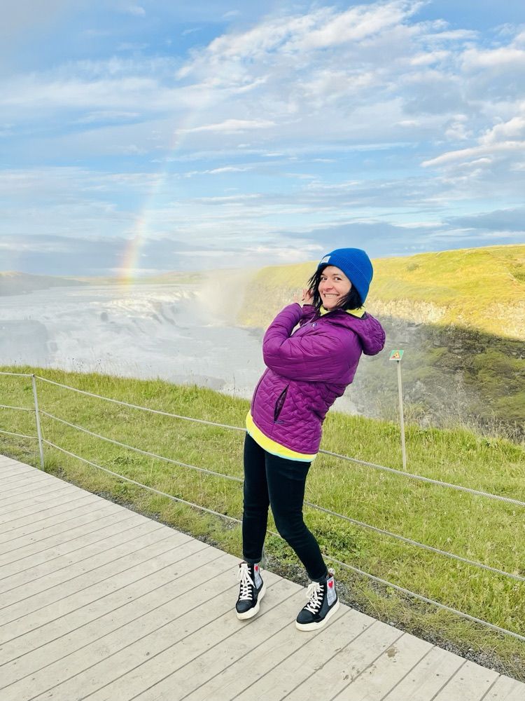 Anne steht auf dem Holzsteg über dem Wasserfall Gullfoss, hinter ihr ein Regenbogen
