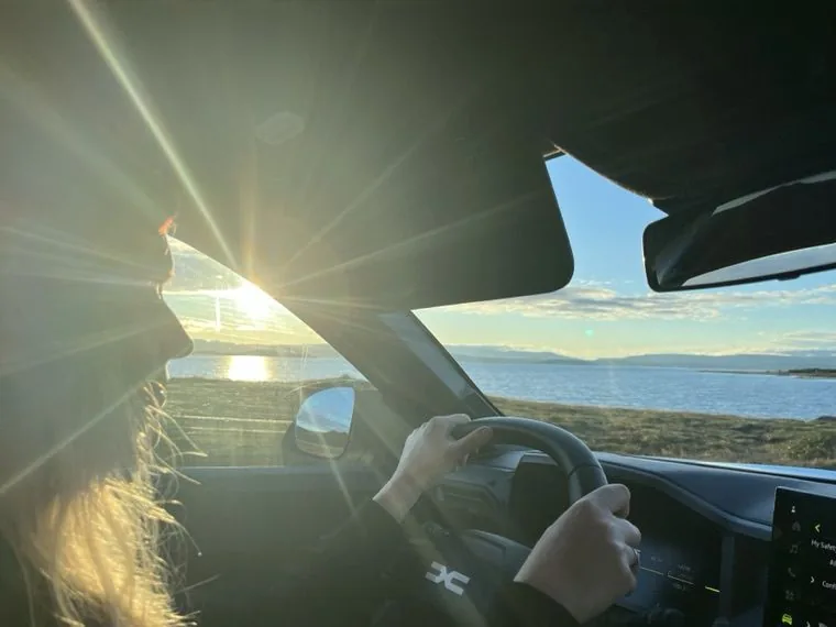 Blick aus dem Auto während der Fahrt: die Fahrerin am Lenkrad, vor der Windschutzscheibe tiefstehende Sonne über einer Bucht