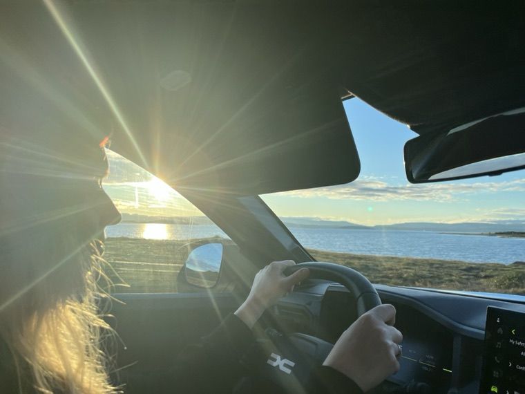 Blick aus dem Auto während der Fahrt: die Fahrerin am Lenkrad, vor der Windschutzscheibe tiefstehende Sonne über einer Bucht